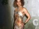 Halle Berry posing at a red carpet event wearing a shimmering gold halter gown with metallic gradient detailing, chunky gold cuff bracelet, and short curly bob hairstyle, standing in front of a green textured backdrop.