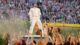 Bad Bunny performing at the Super Bowl LX Halftime Show. He is wearing a full-white Zara combination. Bad Bunny is standing on the top of a white truck, which is surrounded by dancers.